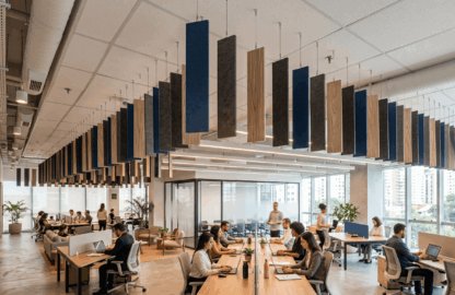 Um escritório corporativo moderno e amplo com dezenas de baffles acústicos Brazil Acoustic suspensos no teto. Os painéis de PET Acústica, em tons de azul, cinza e textura amadeirada, flutuam sobre as estações de trabalho onde colaboradores estão focados em seus laptops. Janelas grandes revelam uma vista de São Paulo ao fundo.
