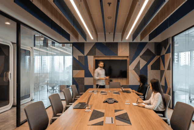 Sala de reuniões corporativa moderna com revestimento geométrico de painel acústico nas paredes em tons de azul e cinza, teto equipado com baffles e iluminação em LED, e profissionais ao redor de uma mesa de madeira.
